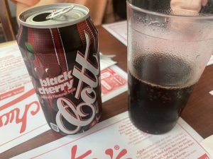 can of Cott black cherry soda next to a glass half filled with it on a table at Schwartz's Deli
